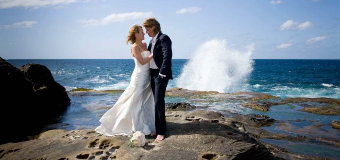 Summer wedding themes a couple stands on a rock in their wedding clothes while waves break behind them.